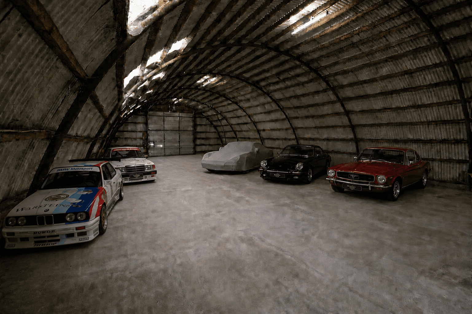 Classic race and sports cars parked inside a large, arched corrugated metal hangar with sunlight.