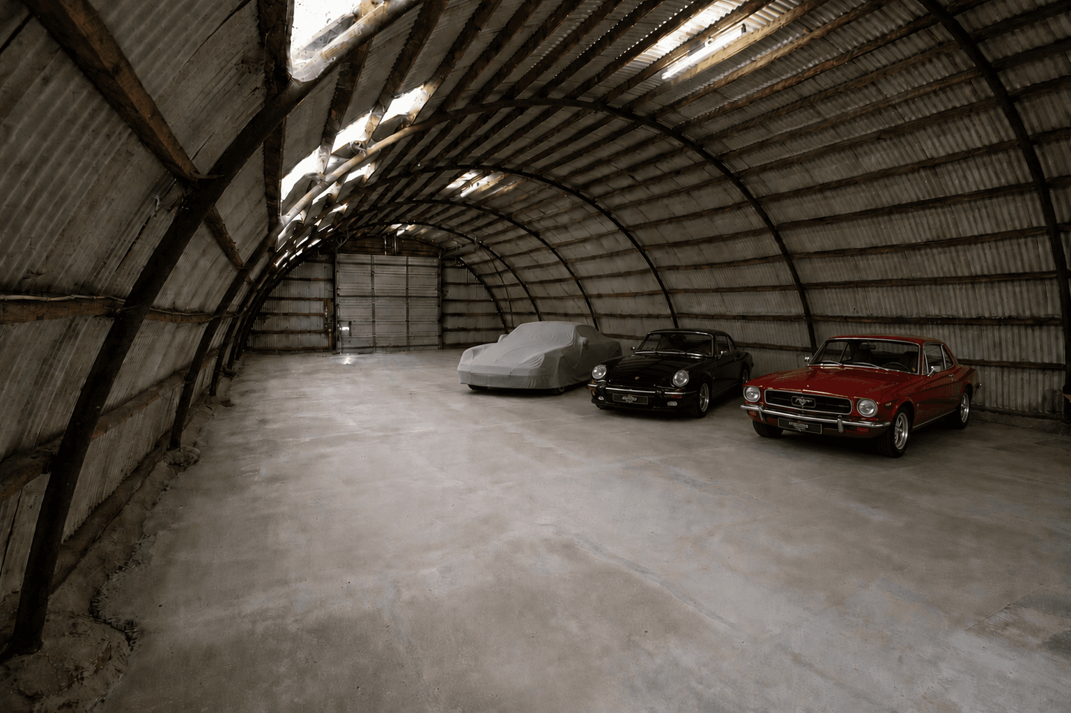 Classic red Mustang and black Porsche parked inside a large, arched corrugated metal hangar.